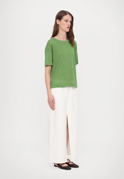 Top verde a maniche corte in tessuto leggero, abbinato a una gonna maxi bianca con spacco frontale e scarpe nere basse.