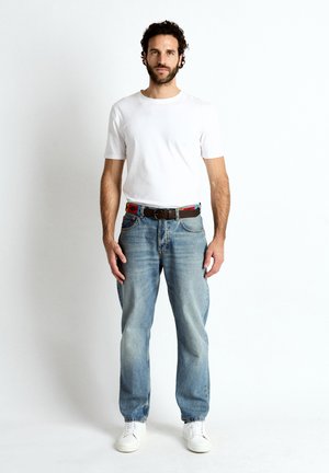 Mężczyzna o ciemnych kręconych włosach i brodzie stoi, ubrany w białą koszulkę, wyblakłe niebieskie dżinsy, kolorowy pasek i białe trampki na białym tle.