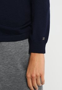 Pull en tricot bleu marine avec un col rond, des poignets côtelés et un petit logo doré près du poignet, assorti à un pantalon en tissu gris.
