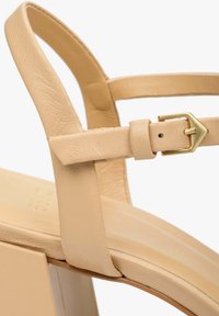 Beżowe skórzane sandały na wysokim obcasie z paskiem z metalowym zapięciem. But ma gładką teksturę i kwadratowy wzór obcasa.