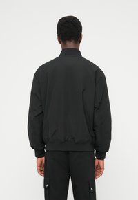 Μαύρο μπουφάν μπομπέρ με όρθιο γιακά, ελαστικά μανίκια και ζώνη στη μέση. Διαθέτει λεία, ματ υφή και μινιμαλιστικό σχεδιασμό.