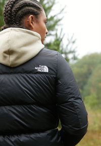 Black puffer jacket with glossy texture, featuring white logo on the left shoulder, worn over a beige hooded sweatshirt.
