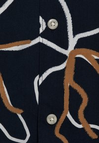 Námořnická látka s abstraktními hnědými a bílými linkami. Obsahuje bílý knoflík na léze, který zdůrazňuje hladkou strukturu a detaily vzoru.