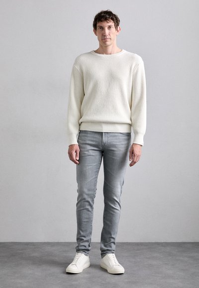 White ribbed knit sweater with long sleeves, paired with light gray slim-fit jeans and white sneakers, standing on a neutral background.