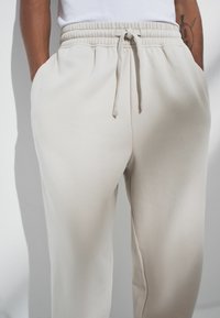 Gai bēšas treniņbikses ar elastīgu jostasvietu un auklu. Iezīmētas ar sānu kabatām un mīkstu, gludu tekstūru.