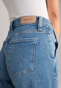 Blue denim jeans with a high waist, single back pocket, and a tan leather label reading "JOOP!" on the waistband.