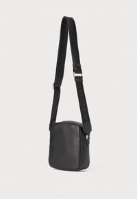 Borsa a tracolla in pelle nera con una texture liscia, forma quadrata, tracolla regolabile con logo e chiusura superiore con zip.