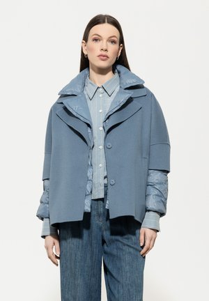 Blue layered jacket with a puffy collar and sleeves, featuring a smooth texture. Worn over a light blue striped shirt and denim pants.