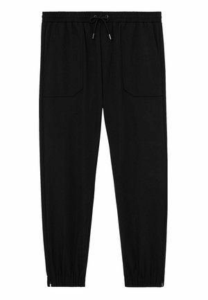 Persona che indossa pantaloni con elastico in vita neri e una giacca scura con trama, con la mano destra nella tasca dei pantaloni, appoggiata a un muro bianco.