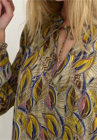 Multifarvet bluse med et bladmønster i gule, blå og brune nuancer. Lavet af letvægtsstof med en nøglehulsudskæring og bindebånd.