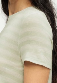 Κοντινό πλάνο ενός ατόμου που φορά ένα πλεκτό κοντομάνικο τοπ με ανοιχτό πράσινες και λευκές ρίγες, με ορατά σκοτεινά μακριά μαλλιά.