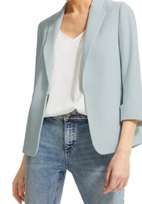 Ljusblå kavaj med en slät yta, öppen front och trekvartslånga ärmar, i kombination med en vit topp och jeans.