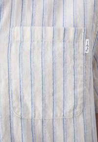 Gestreept shirt in lichtcrème met dunne blauwe lijnen, met een patchzak en een klein merkembleem nabij de naad. Zachte stoftextuur.