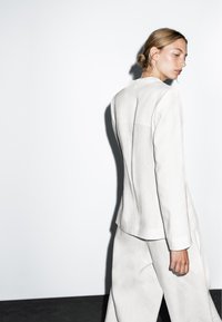 Donna con capelli biondi raccolti, che indossa un abito bianco di taglio morbido, mentre cammina e guarda di lato su uno sfondo bianco semplice.