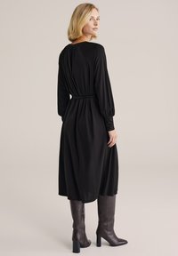 Fekete hosszú ujjú ruha, testhezálló derékkal, puha tapintású anyagból, középhosszú szoknyával. Barna magas sarkú csizmával párosítva.