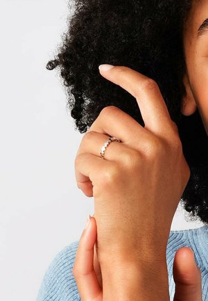 Anello in oro rosa con un design delicato, caratterizzato da piccoli elementi rotondi disposti uniformemente attorno all'anello, indossato su un dito sopra un tessuto testurizzato.