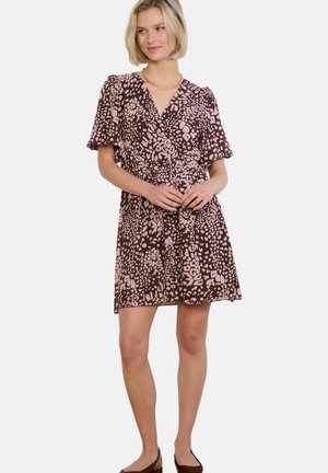 Ženska stoji oblečena v kratko rjavo obleko z roza abstraktnimi lisami, kratkimi rokavi, v-izrezom in pasno vrvico, kombinirano s črnimi balerinkami.