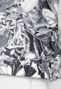 Černobílé sportovní tričko s květinovým vzorem a měkkou texturou, s výrazným logem Nike poblíž lemu a detailními bočními rozparky.