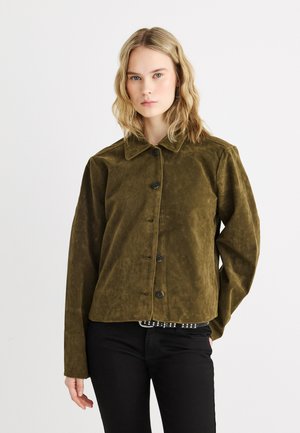 Mladá žena s vlnitými blond vlasy, oblečená v olivově zelené semišové bundě na knoflíky a černých kalhotách, stojící před jednobarevným bílým pozadím.