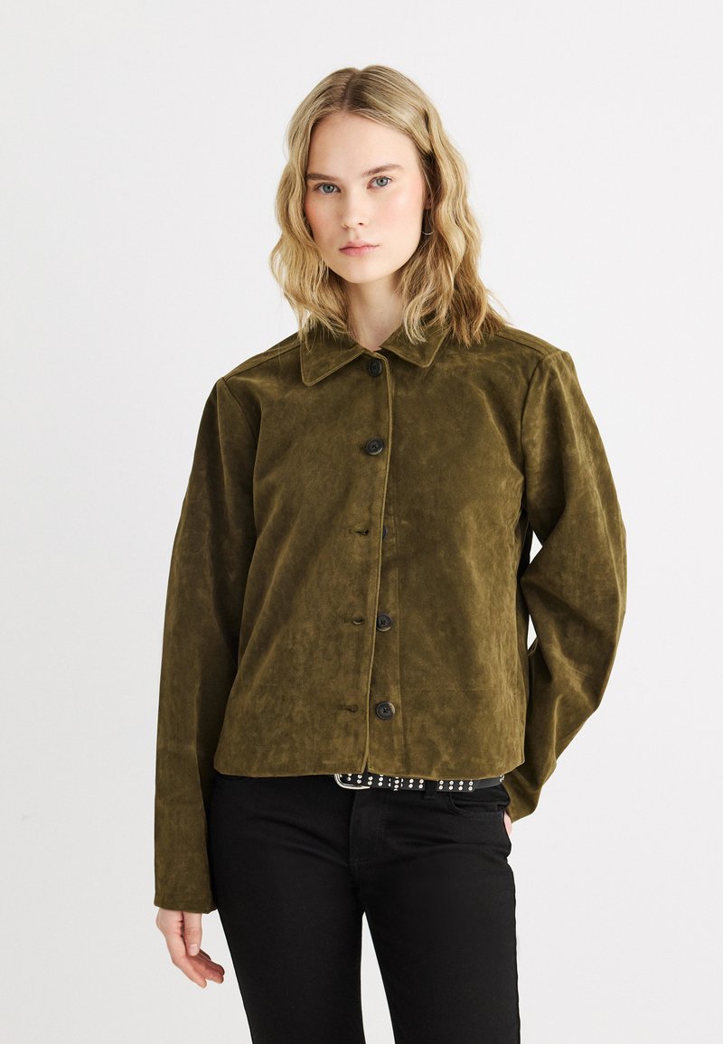 Νεαρή γυναίκα με ξανθά κυματιστά μαλλιά, που φοράει πράσινο ελαιώδες σουέντ σακάκι με κουμπιά και μαύρο παντελόνι, μπροστά σε απλό λευκό φόντο.