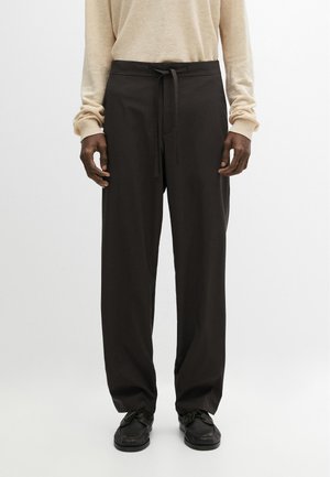 Mann trägt lockere schwarze Hosen mit Kordelzug in der Taille und schwarze Schuhe, kombiniert mit einem beigen Langarmtop, stehend vor einem einfarbigen Hintergrund.