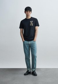 Μαύρο κοντομάνικο μπλουζάκι με γραφικό αρκούδας στο στήθος, συνδυασμένο με ανοιχτό μπλε τζιν με γυρισμένα μανίκια και μαύρα παπούτσια σε ανοιχτό φόντο.