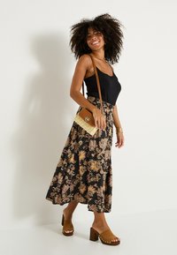 Haut sans manches noir associé à une longue jupe à motif floral noir, or et rose, complété par des mules à plateforme beiges et un sac en osier.