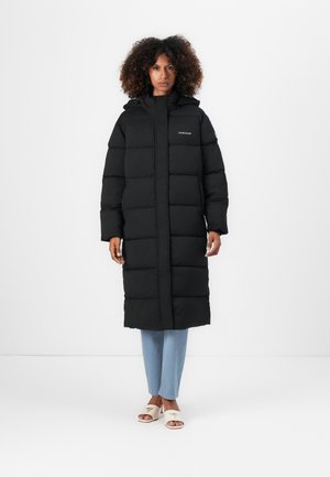 Langer schwarzer Pufferjacke mit Kapuze, ausgestattet mit einem gesteppte Design und einem Reißverschluss auf der Vorderseite. Kombiniert mit hellblauen Hosen und hellen Sandalen.