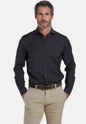 Man met kort zout-en-peper haar, draagt een zwart overhemd met knopen en beige broek met een bruine riem, handen in de zakken, neutrale achtergrond.