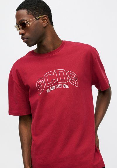 Crvena pamučna majica s bijelim logotipom "GCDS MILANO ITALY 1988" u podebljanom fontu. Kroj je opušten, s kratkim rukavima i okruglim izrezom.