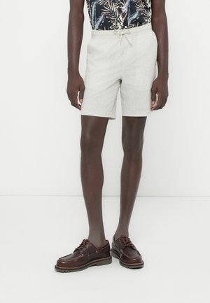 Pantaloncini in lino grigio chiaro con vita con coulisse, abbinati a una camicia scura a fantasia e mocassini da barca in pelle marrone.