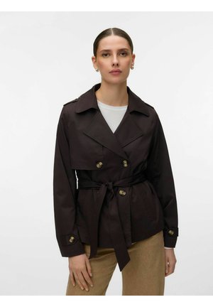 Femme aux cheveux tirés en arrière portant un trench-coat marron foncé ceinturé sur un haut blanc et un pantalon beige, debout devant un fond uni.