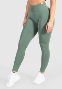 Leggings verdi a vita alta con una texture liscia e un design aderente. Abbinati a scarpe da ginnastica bianche e una maglietta nera.