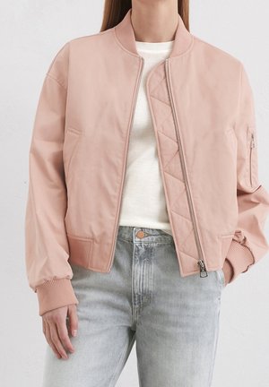 Frau trägt eine hellrosa Bomberjacke über einem weißen Pullover und hellblauen, hoch taillierten Jeans, steht mit einer Hand in der Tasche.