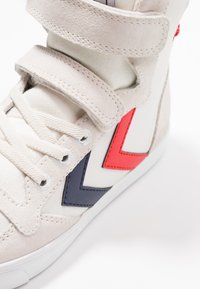 Sko av tyg i vitt och grå mocka. Har röda och bl å chevron-accenter, kardborreband och traditionell snörning.