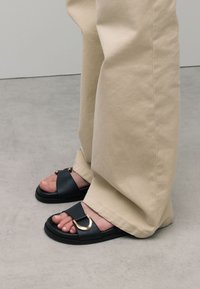 Sandales en cuir noir à bout ouvert, avec un accent de quincaillerie circulaire dorée et une semelle noire épaisse, associées à un pantalon beige clair.