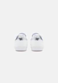 Bílé tenisky s hladkým koženým svrškem a stříbrnými třpytivými detaily na patě. Texturovaná gumová podrážka a minimalistický design.