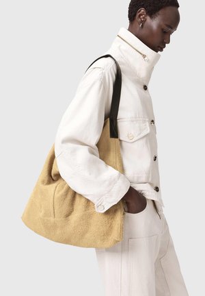 Beige terry-kangaskassi, jossa rennon pehmeä muotoilu ja mustat nahkahihnat. Malli pukeutunut valkoiseen takkiin, jossa näyttävät napit ja korkea kaulus.