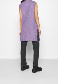 Ärmellose lavendelfarbene Strick-Tunika mit seitlichen Schlitzen, kombiniert mit schwarzen Denim-Hosen und klobigen schwarzen Stiefeln, vor neutralem Hintergrund.
