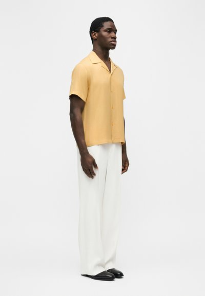 Άνδρας που στέκεται με κίτρινο κοντομάνικο πουκάμισο με κουμπιά, λευκό παντελόνι φαρδιάς γραμμής και μαύρα παπούτσια, μπροστά σε απλό φωτεινό φόντο.