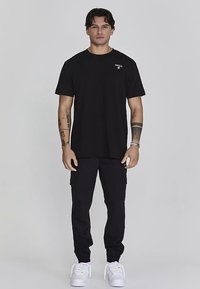 Črna majica z okroglim izrezom z majhnim logotipom na prsih, kombinirana s črnimi cargo hlačami in belimi supergami, vse v nevtralnem okolju.