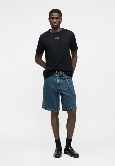 Νεαρός άνδρας στέκεται με τα χέρια στην πλάτη, φοράει μαύρο t-shirt GCDS, μπλε τζιν σορτς με ζώνη και αλυσίδα, μαύρες κάλτσες και μαύρα παπούτσια.
