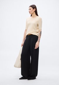 Top bej tricotat cu mâneci scurte, asortat cu pantaloni negri cu picior larg. Modelul ține o geantă crem și are un tatuaj subtil pe brațul stâng.