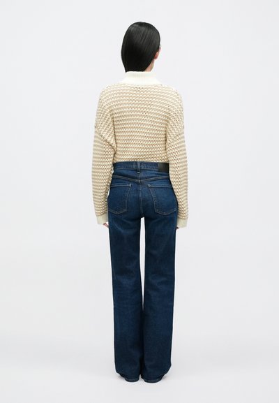 Krémový a béžový pletený sveter s vysokým golierom, v kombinácii s tmavomodrými džínsami širokých nohavíc s vreckami na zadnej strane a pásom so značkou.