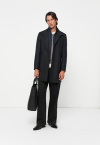 Manteau en laine noir avec un col structuré, superposé sur une chemise à motifs. Un pantalon noir et un sac fourre-tout noir élégant complètent l'ensemble.