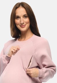 Felpa rosa chiaro con colletto rotondo, dettaglio con tasca laterale con cerniera e polsini a coste. Tessuto morbido con vestibilità rilassata.