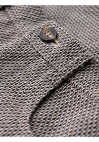 Gros plan d’un tissu tissé marron et beige texturé avec un seul bouton marron foncé cousu sur un rabat plié.