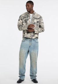 Camouflagejack in beige en grijs, met twee borstzakken. Draag het met lichtblauwe distressed jeans en zwarte schoenen.