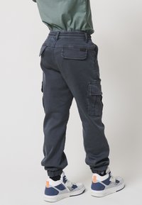 Mornarske cargo hlače z elastiko v pasu, zategnjenimi gležnji in več stranskimi žepki, v kombinaciji z belimi in modrimi teniskami z oranžnimi detajli.