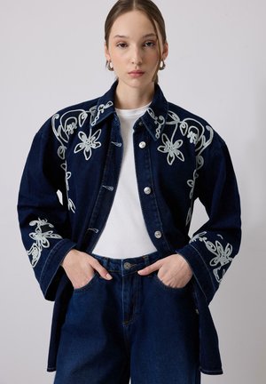 Jeune femme portant une veste en denim bleu foncé brodée sur une chemise blanche et un jean bleu foncé, mains dans les poches, fond neutre.